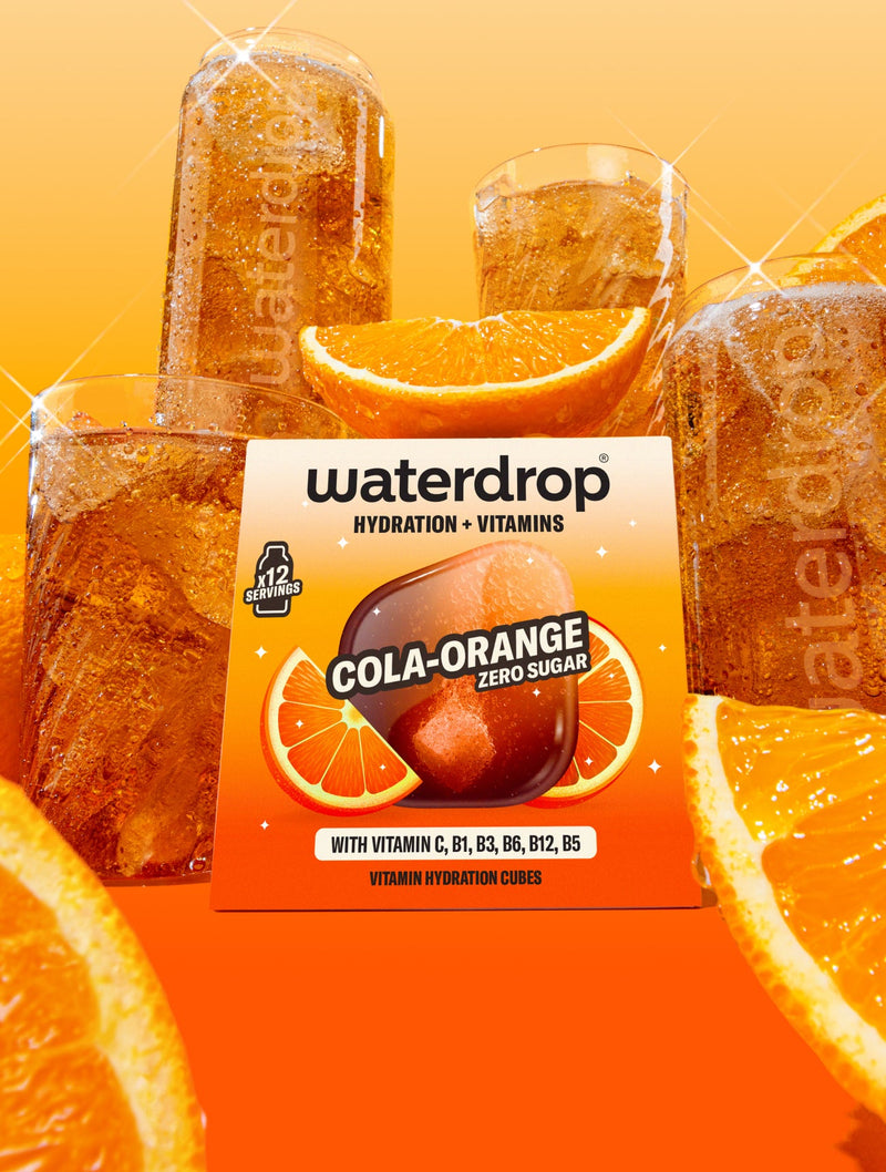 Krabička príchute Cola Orange s 12 ks umiestnená v strede, v pozadí sú poháre waterdrop® naplnené nápojom a okolo sú zobrazené kúsky pomaranča.