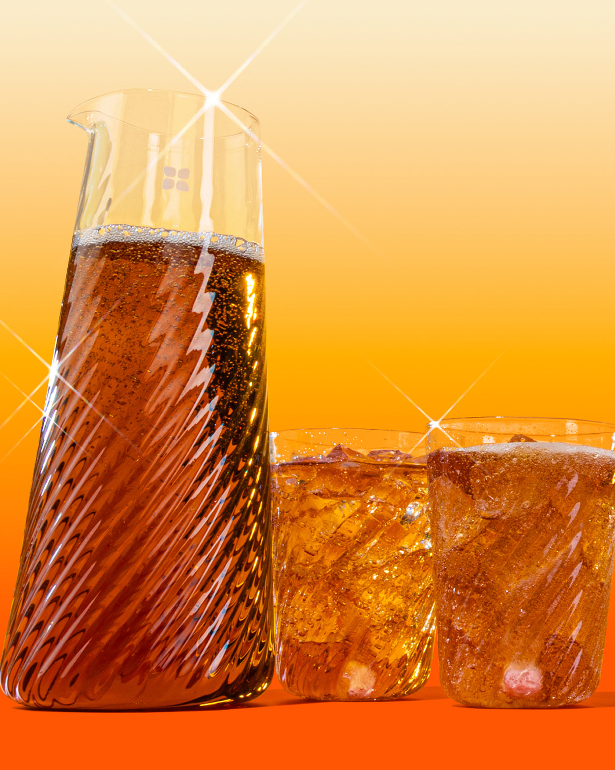Sklenená karafa a dva vlnité poháre naplnené tmavým perlivým nápojom s ľadom, umiestnené na oranžovom pozadí.