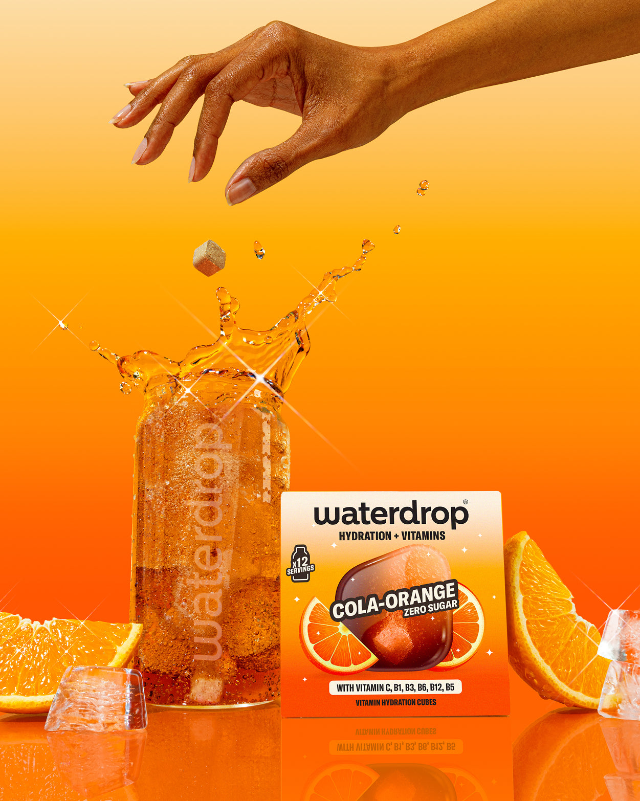 Ruka púšťa kocku Waterdrop Cola Orange do pohára s perlivým nápojom, vedľa krabičky príchute, plátkov pomaranča a ľadu, na oranžovom pozadí.