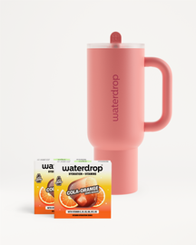 Ružový termohrnček Waterdrop s viečkom a slamkou umiestnený vedľa dvoch krabičiek príchute Cola Orange na bielom pozadí.