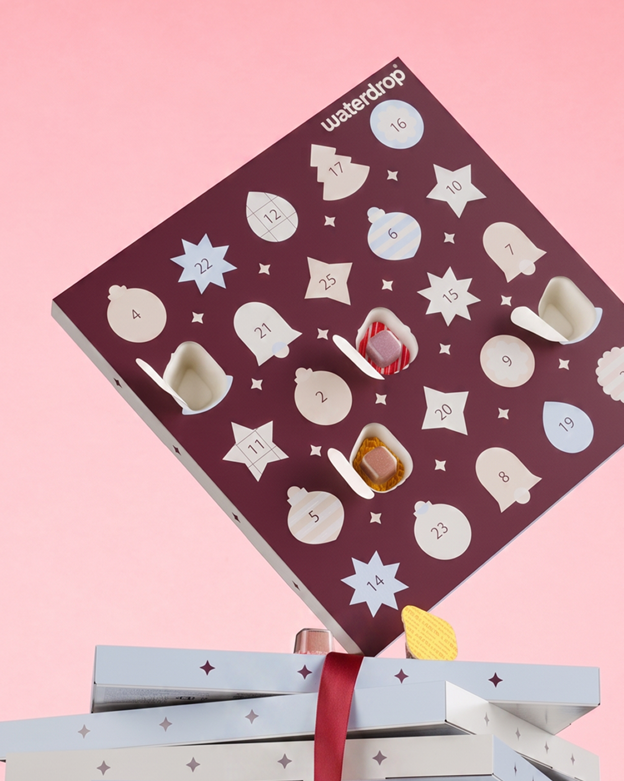 Purpurový adventný kalendár so hviezdičkovými dverami na ružovom pozadí, nad dvoma zabalenými darčekmi.