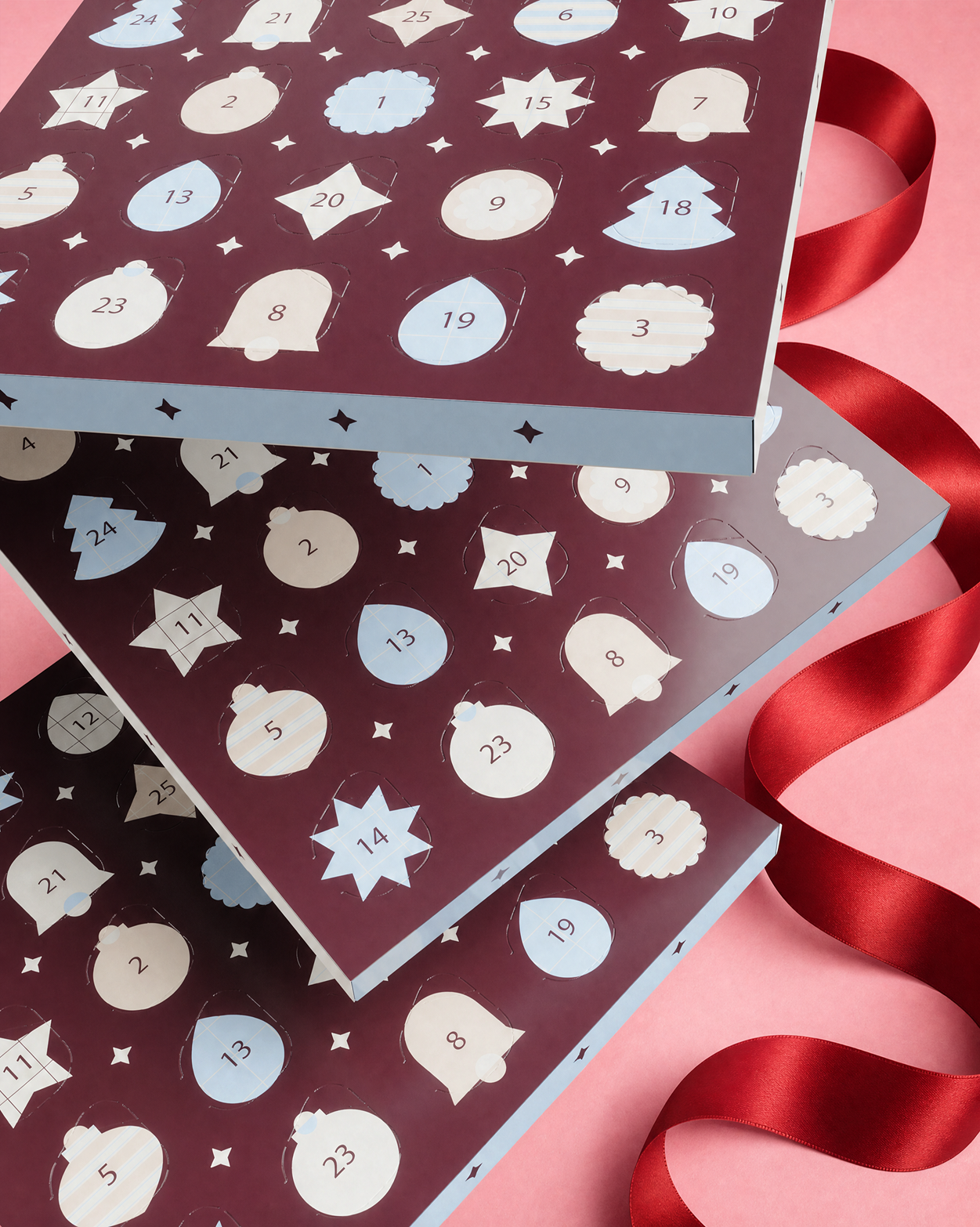 Tri poskladané Advent kalendárové krabičky s číslovanými dvierkami a dekoratívnymi tvarmi, zviazané červenou stuhou na ružovom pozadí.