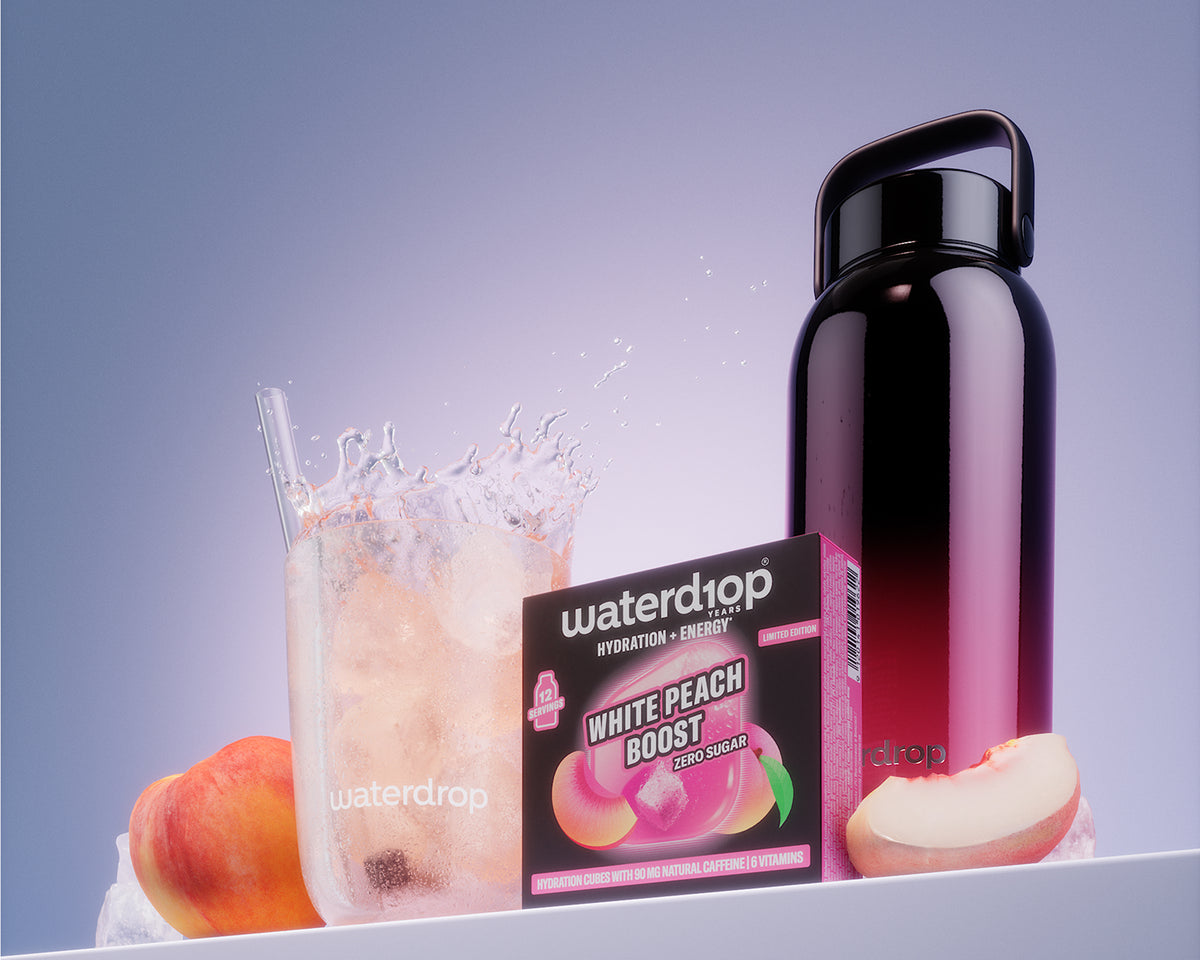 Box produktu White Peach Boost vedľa pohára s ľadom a čiernej opakovane použiteľnej fľaše na bielom regále.