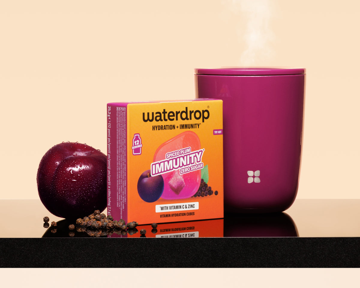 Fialová šálka s parou pri oranžovom balení Waterdrop, slivka a roztrúsené semienka na lesklom povrchu.