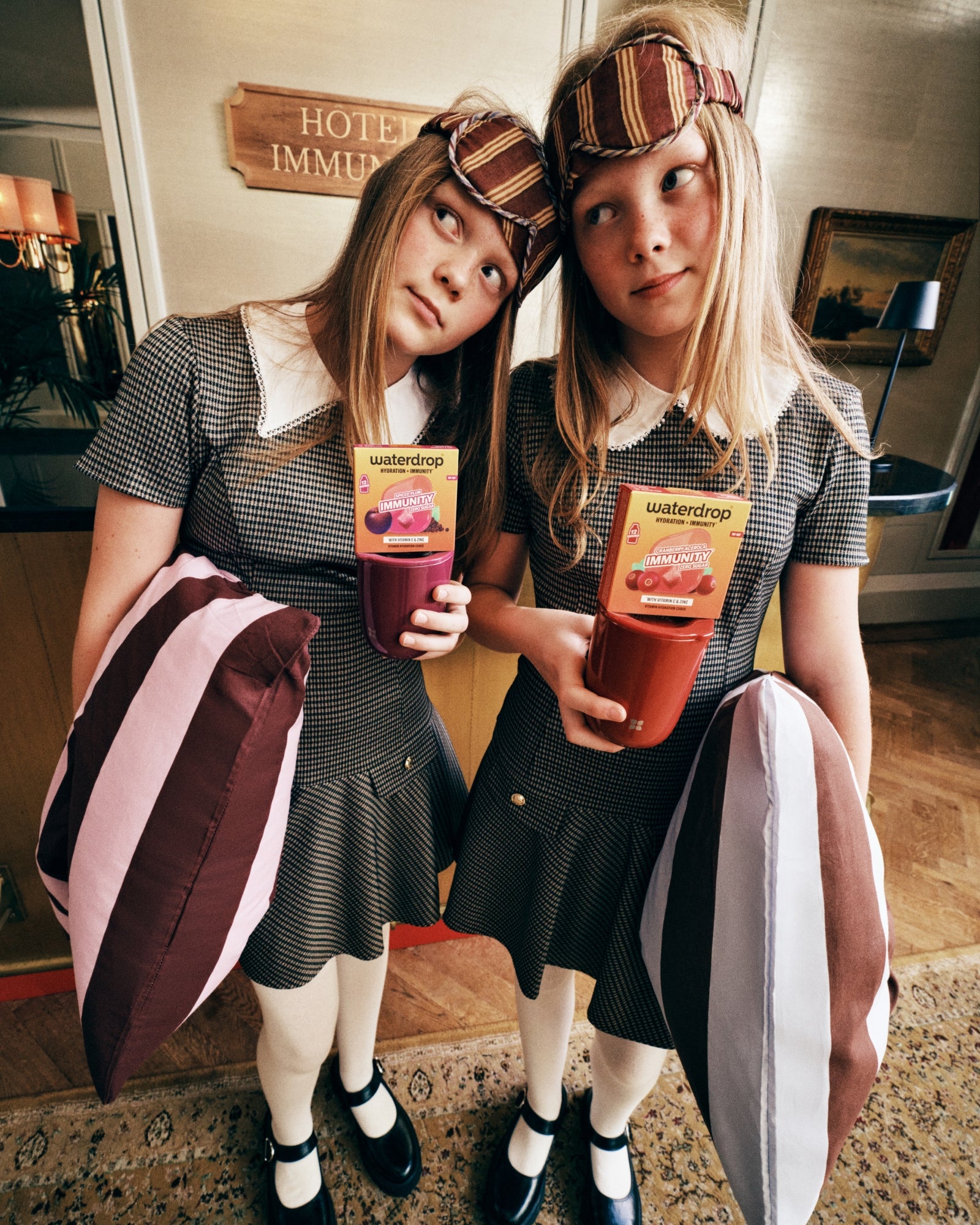 Dve dievčatá v kocovaných šatách s pruhovanými čelenkami stoja bok po boku v hotelovom vestibule, každá drží vrecko so šťavou.